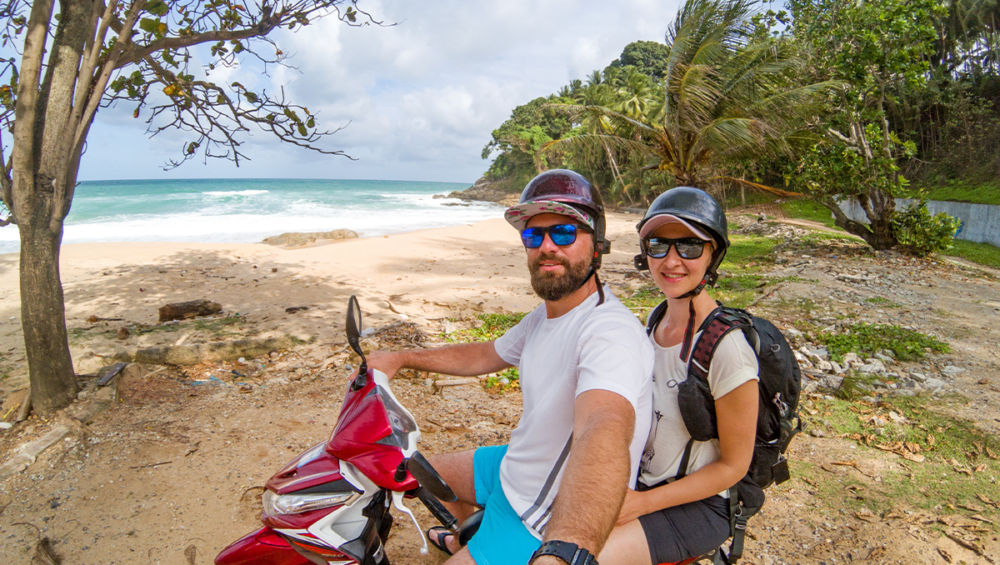 A smiling tourist couple wearing sunglasses and helmets on a motorbike, looking at the camera, standing beside a beach in Phuket, ready to explore the island by scooter.