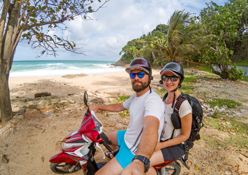 A smiling tourist couple wearing sunglasses and helmets on a motorbike, looking at the camera, standing beside a beach in Phuket, ready to explore the island by scooter.